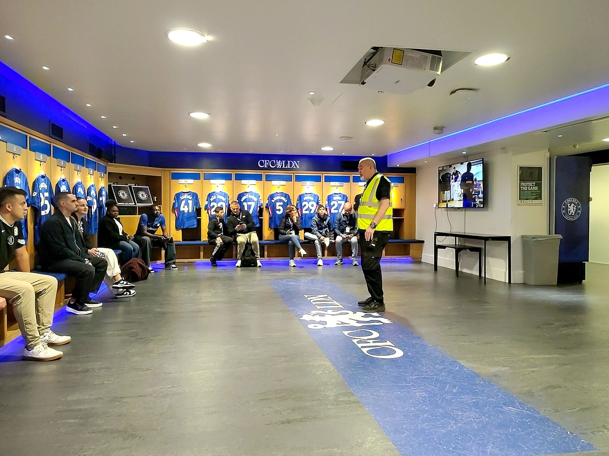 Grupa zwiedzających słucha przewodnika w szatni Chelsea FC, otoczonej klubowymi koszulkami zawodników.