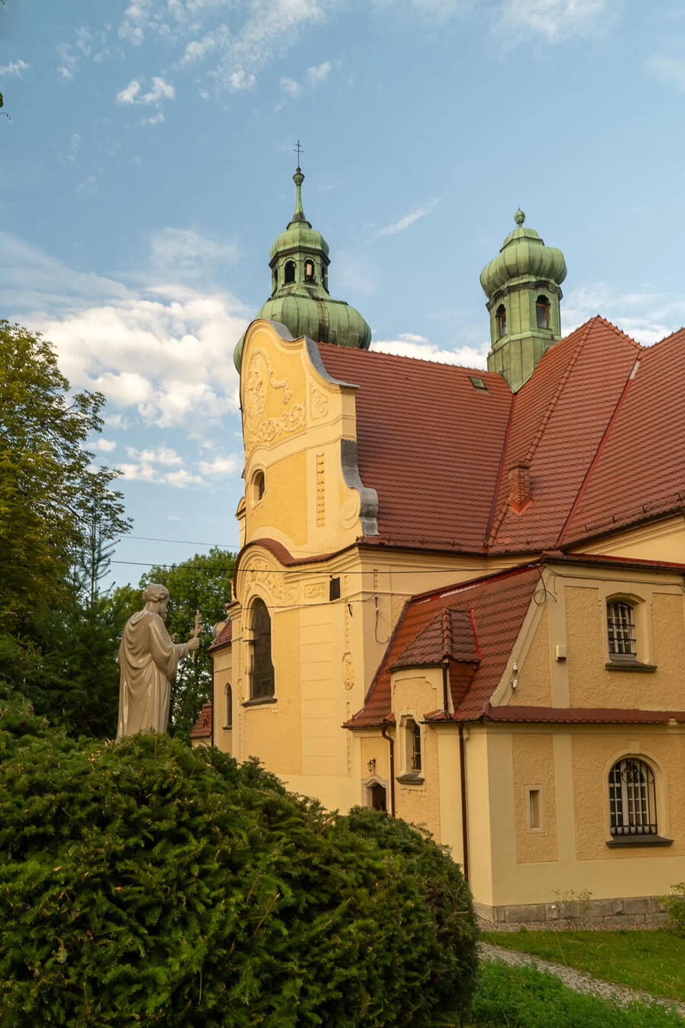 Kościół Wniebowzięcia NMP w Polanicy Zdrój — widok od strony absydy z brukowaną alejką i spacerowiczami wśród drzew