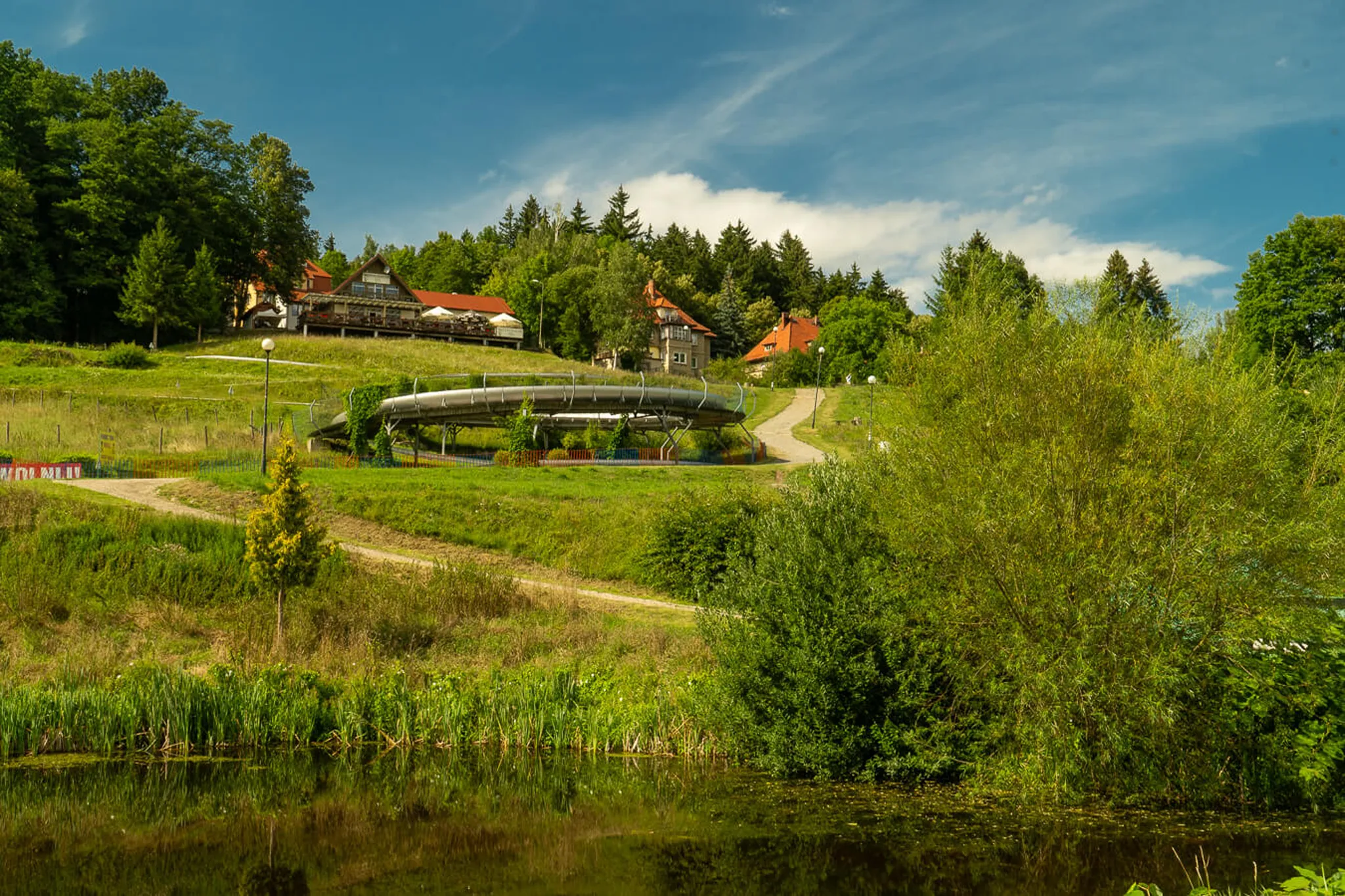 Letni tor saneczkowy Góralka w Polanicy Zdrój — spiralna stalowa rynna toru na zielonym zboczu ze stawem na pierwszym planie